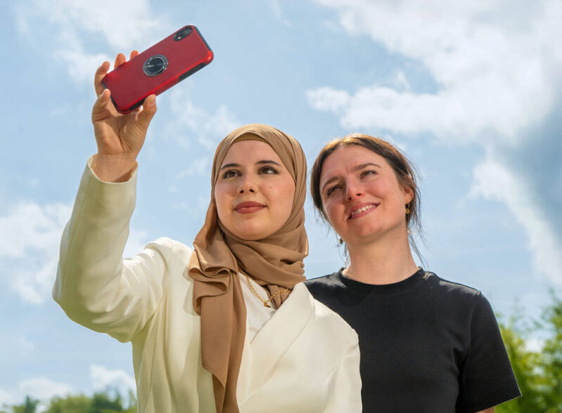 two females, one wearing a head scarf, smiling taking a selfie together