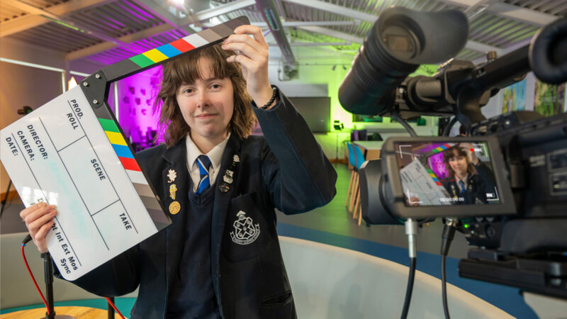 female school student holds clapper in front of TV camera in media suite